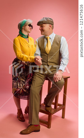 Mature woman smiles while standing beside man seated on wooden stool, dressed in brown vest and trousers. Both posing elegantly against pink studio background. 121475616