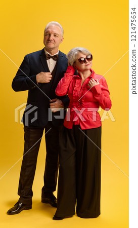 Confident man in formal ensemble gazing forward standing with woman in striking red blouse, both radiating elegance, against bright yellow background. Confident man in formal ensemble gazing forward standing with woman in striking red blouse, both radiating elegance, against bright yellow background. 121475654