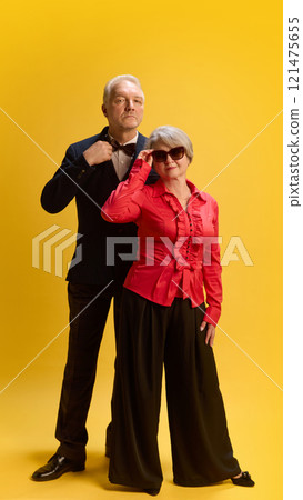 Sophisticated pair, man in tailored suit standing beside woman in modern attire and shades, posing confidently against yellow studio background. 121475655