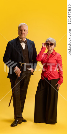 Mature couple, man in plaid blazer and scarf with woman in cream jacket and sunglasses, posing arm-in-arm against blue studio background. 121475659