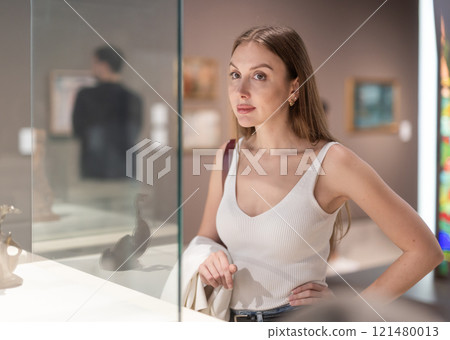 Young female visitor stands near glass display case and examines valuable exhibit 121480013