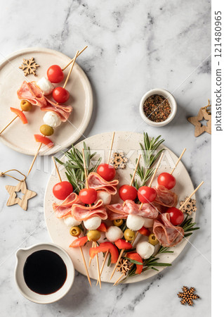 Skewers with mozzarella, ham, olives, peppers, tomatoes. Festive canapes or antipasti on white tray for new year table, top view 121480965