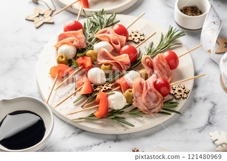 Snack skewers with mozzarella, ham, olives, peppers, tomatoes. Festive canapes or antipasti on white tray for new year table 121480969