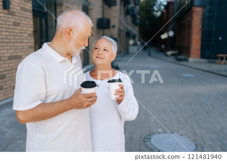 Portrait of elderly man and woman standing on urban paved street, holding coffee cups and loving looking at each other. Happy senior couple enjoying sunny day together during summer walking. 121481940