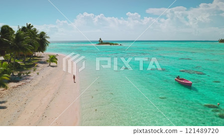 Aerial view of a tourist walking along a beautiful pink sand beach on a tropical island in aitutaki, cook islands, with a small boat anchored in turquoise water 121489720