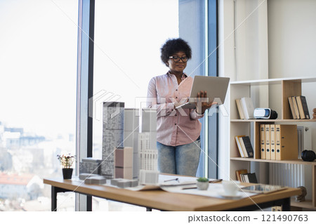 African American female architect in casual attire develops building project using laptop. Bright workspace with architectural models and natural light enhances creative process. 121490763