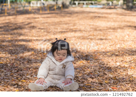[One-year-old child sitting and playing in a winter park covered with fallen leaves] 121492762