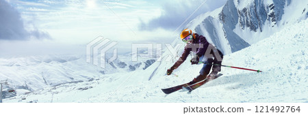 Skier making sharp turn on snowy mountain slope, scattering snow, with dramatic clouds and expansive snowy peaks in distance. 121492764