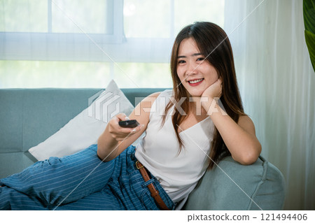 Happy female fun comedy video holding remote watching television, Asian young woman smiling sitting relaxing watch TV holding remote control on sofa in living room, Activity lifestyles concept Happy female fun comedy video holding remote watching television, Asian young woman smiling sitting relaxing watch TV holding remote control on sofa in living room, Activity lifestyles concept 121494406