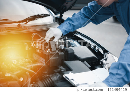Auto technician in a workshop writing on a clipboard checking car's engine for maintenance. Mechanic expertise focusing on safety and care. Business in automotive industry smiling while working. Auto technician in a workshop writing on a clipboard checking car's engine for maintenance. Mechanic expertise focusing on safety and care. Business in automotive industry smiling while working. 121494429