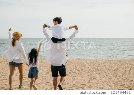 At a sunset sea beach happy family of four stands with raised arms. The father carries his son on shoulders epitomizing the joy and togetherness of a carefree beach vacation. Family on beach vacation 121494451