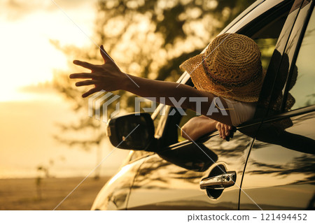 Woman wearing a stylish hat sits in front seat waving hand out car window at sunset feeling the wind. Happy weekend travel enjoy summer at beach. road trip and freedom concept 121494452