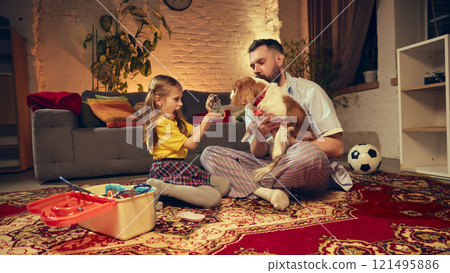 Girl playfully using a toy dental impression, playing with her father and their dog in cozy and warm living room in the evening 121495886