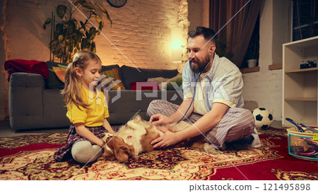 Preschool girl with her father attentively caring for their dog, using stethoscope and other toy tools, sitting on vibrant carpet in cozy living room. 121495898