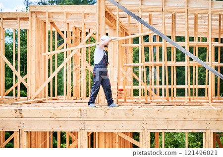 Carpenter constructing wooden-framed house near. Man holds large beam on his shoulder and is dressed in work clothes and helmet. Modern and environmentally-friendly construction concept. Carpenter constructing wooden-framed house near. Man holds large beam on his shoulder and is dressed in work clothes and helmet. Modern and environmentally-friendly construction concept. 121496021