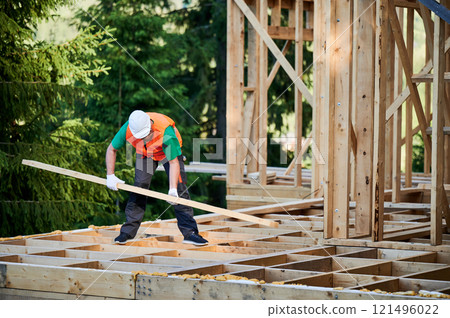 Carpenter constructing wooden-framed house near the woods at sunset. Man holding truss and dressed in work clothes and helmet. Concept of modern and environmentally-friendly construction. 121496022