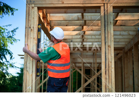 Carpenter constructing two-storey wooden frame house. Man inspects walls for levelness using spirit level, wearing protective overalls, helmet and orange vest. Concept of contemporary construction. Carpenter constructing two-storey wooden frame house. Man inspects walls for levelness using spirit level, wearing protective overalls, helmet and orange vest. Concept of contemporary construction. 121496023