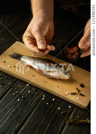 A person carefully seasons a freshly caught fish with aromatic spices and herbs on a wooden cutting board, evoking a cozy culinary atmosphere in a rustic kitchen. A person carefully seasons a freshly caught fish with aromatic spices and herbs on a wooden cutting board, evoking a cozy culinary atmosphere in a rustic kitchen. 121496381