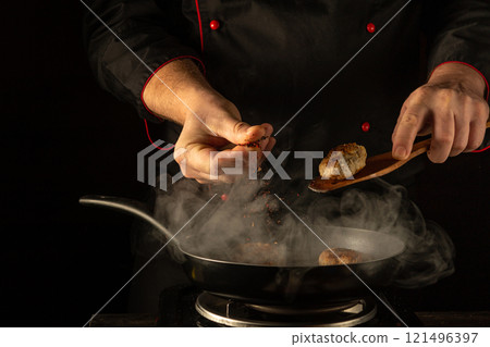 In a dimly lit kitchen, a skilled chef crafts delicious meatballs, skillfully seasoning them while steam rises from the pan. The focus on his precise movements showcases culinary artistry 121496397