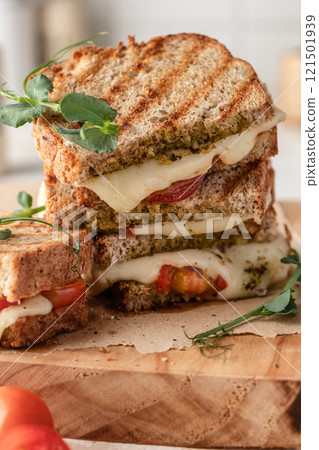 Grilled sandwich panini with mozzarella cheese, cherry tomatoes and pesto on wooden board, kitchen table background close up. Traditional italian food Grilled sandwich panini with mozzarella cheese, cherry tomatoes and pesto on wooden board, kitchen table background close up. Traditional italian food 121501939