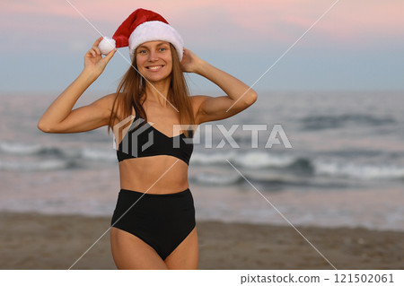 Attractive smiling young woman wearing Santa Claus hat and black bikini on seashore looks at camera. Christmas and New year vacations. Celebrating winter holidays abroad at warm tropical destination. Attractive smiling young woman wearing Santa Claus hat and black bikini on seashore looks at camera. Christmas and New year vacations. Celebrating winter holidays abroad at warm tropical destination. 121502061