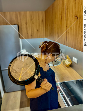 A young girl in the kitchen near an electric stove holds a frying pan in her hands and tries to figure out the equipment 121502660