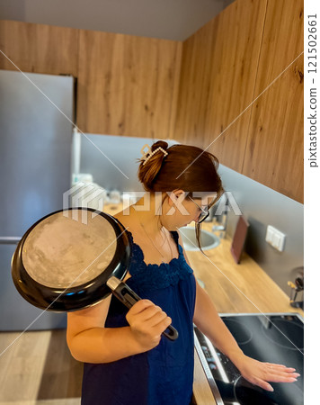 A young girl in the kitchen near an electric stove holds a frying pan in her hands and tries to figure out the equipment 121502661