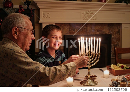 Side view portrait of smiling Jewish boy with grandfather lighting menorah candles during Hanukkah celebration in cozy home setting copy space 121502706