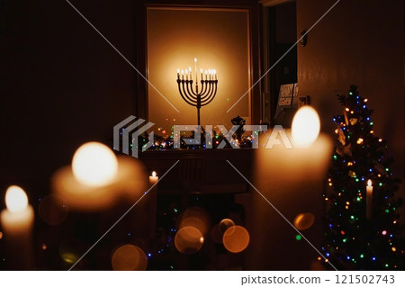 Background image of menorah candles on mantlepiece glowing brightly in dark room with twinkling Christmas lights at night in cozy home setting copy space 121502743