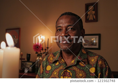 Portrait of adult Black man wearing traditional colorful print and looking at camera surrounded by candles in cozy home setting copy space 121502748