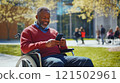 Portrait of grey haired African American man with disability using wheelchair in college campus and holding phone smiling cheerfully copy space 121502961