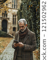 Vertical waist up portrait of senior professor wearing coat and using phone while walking along cobblestone path in classic campus grounds 121502962