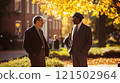 Side view portrait of two professors dressed in formal suits chatting casually standing under autumn trees with red brick university buildings in background copy space 121502964