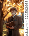 Vertical portrait of classic senior man with white beard wearing vintage suit and hat while reading book in autumn park or college campus 121502968