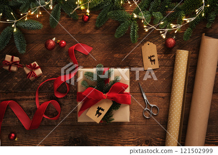 Festive gift wrapping scene with garland lights. Kraft brown paper rolls, red velvet ribbon, labels, scissors and finished wrapped present. 121502999