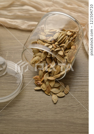 Scattered pumpkin seeds in transparent glass jar on wooden table, beige cloth on the background 121504710