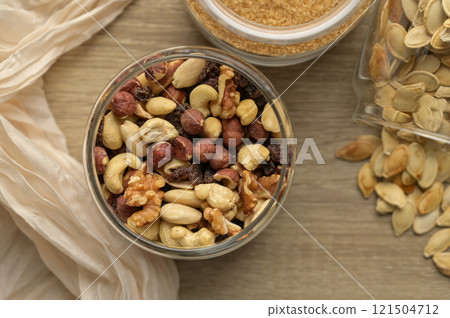 Top view closeup cane brown sugar, nuts mix, raisins and scattered pumpkin seeds in transparent glass jars on wooden table, beige cloth on the background Top view closeup cane brown sugar, nuts mix, raisins and scattered pumpkin seeds in transparent glass jars on wooden table, beige cloth on the background 121504712