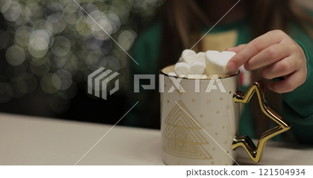 Girl eats marshmallow. Close-up of little girl in a green Christmas sweater drinks cocoa with marshmallows in the kitchen on glowing Christmas tree background. Christmas mood. Girl eats marshmallow. Close-up of little girl in a green Christmas sweater drinks cocoa with marshmallows in the kitchen on glowing Christmas tree background. Christmas mood. 121504934