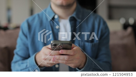 Male typing on smartphone. Texting at phone, sitting on sofa in living room. Surfing internet social media, watching videos. Male typing on smartphone. Texting at phone, sitting on sofa in living room. Surfing internet social media, watching videos. 121504953