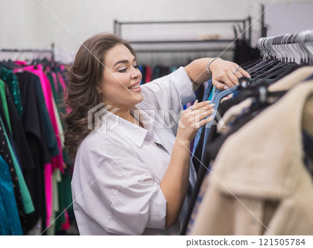 A smiling fat woman in a plus size store chooses clothes while sorting through the hangers. A smiling fat woman in a plus size store chooses clothes while sorting through the hangers. 121505784
