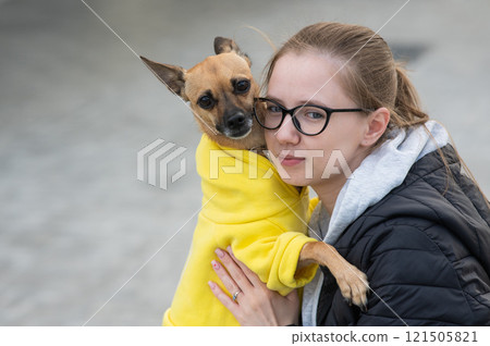 Portrait of a young Caucasian woman hugging a small dog in a jacket.  121505821