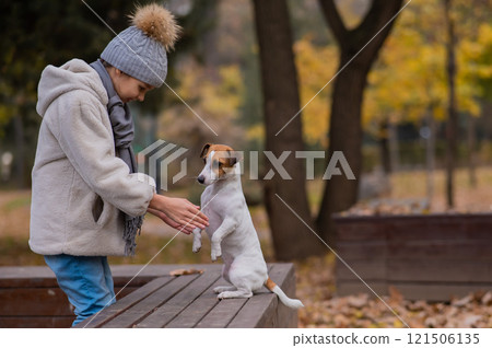 Caucasian girl holding a dog by the paws for a walk in the autumn park. Jack Russell Terrier stands on its hind legs on a bench. 121506135