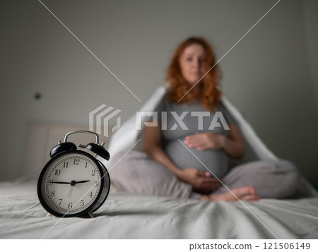 Caucasian pregnant woman sits on the bed and suffers from insomnia. Alarm clock in the foreground. Caucasian pregnant woman sits on the bed and suffers from insomnia. Alarm clock in the foreground. 121506149