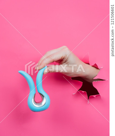 Female hand sticking out of pink paper background with blue curved vibrator sex toy. Vertical photo.  121506601