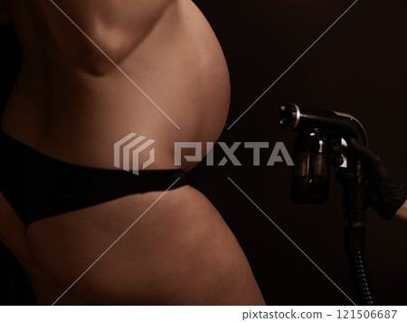 Close-up of the belly of a pregnant woman undergoing an instant tanning procedure. The master applies bronzer with a spray. Close-up of the belly of a pregnant woman undergoing an instant tanning procedure. The master applies bronzer with a spray. 121506687