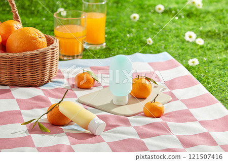 The promotional photo for sunscreen advertisement with picnic set up on green lawn. Two mockup bottle without label displayed in center of pink carpet, fresh orange and juice decorated around The promotional photo for sunscreen advertisement with picnic set up on green lawn. Two mockup bottle without label displayed in center of pink carpet, fresh orange and juice decorated around 121507416