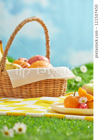 A picnic concept on lush grass with a blue sky backdrop. Frontal view of an empty space for presentation products, next to a dish and a basket filled with some fresh fruits, on a yellow picnic mat. 121507450