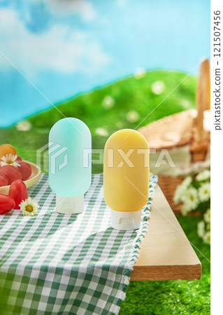 Frontal views of two bottle mockups are shown on a wooden table with a basket, tomatoes, and daisy flowers on a green picnic rug. A concept for a picnic in a lush meadow beneath a blue sky. 121507456