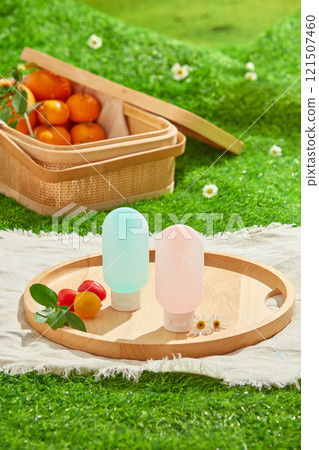 Two bottle mockups are shown in an overhead view on a wooden tray, placed on a white picnic rug, among a tomato, daisy flowers, and an orange basket. Picnic under a blue sky on a lush meadow idea. 121507460