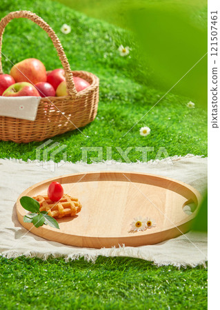 A picnic-themed commercial advertisement, frontal view of an apple basket, dessert, and flowers on green grass, creating an empty wooden tray displaying products. Overhead shot. 121507461